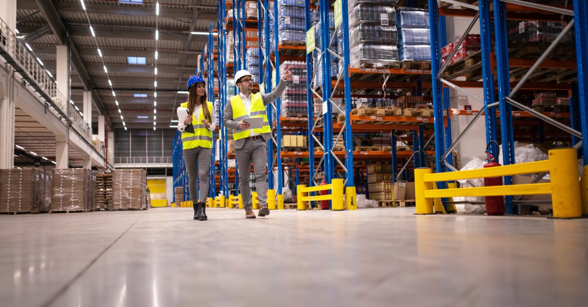 Two warehouse managers wearing high-visibility safety vests, inspecting a large warehouse with full shelves.