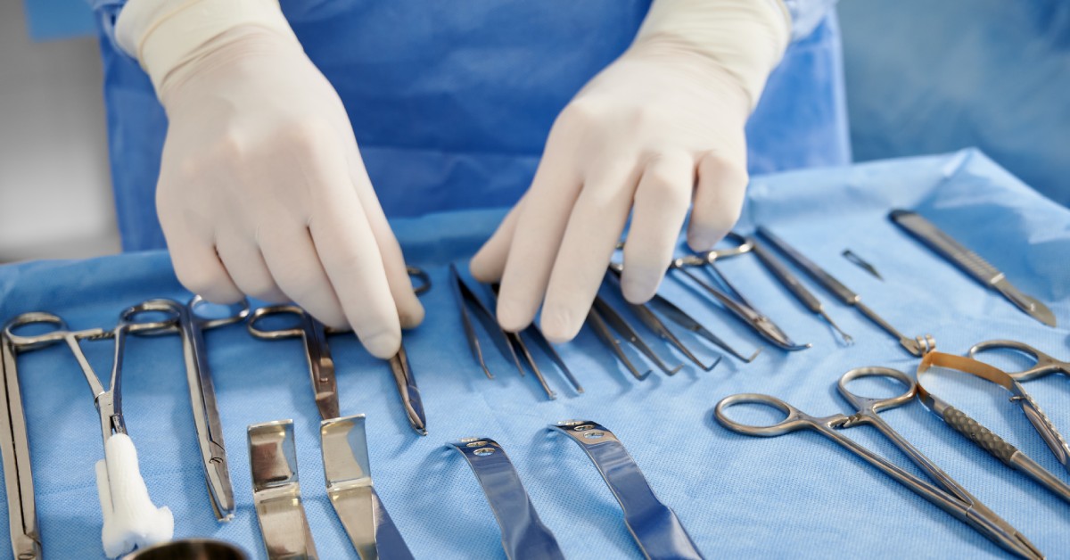 A pair of hands organizes sterilized surgical equipment on a medical tray. There are prepping scissors, forceps, and scalpels.