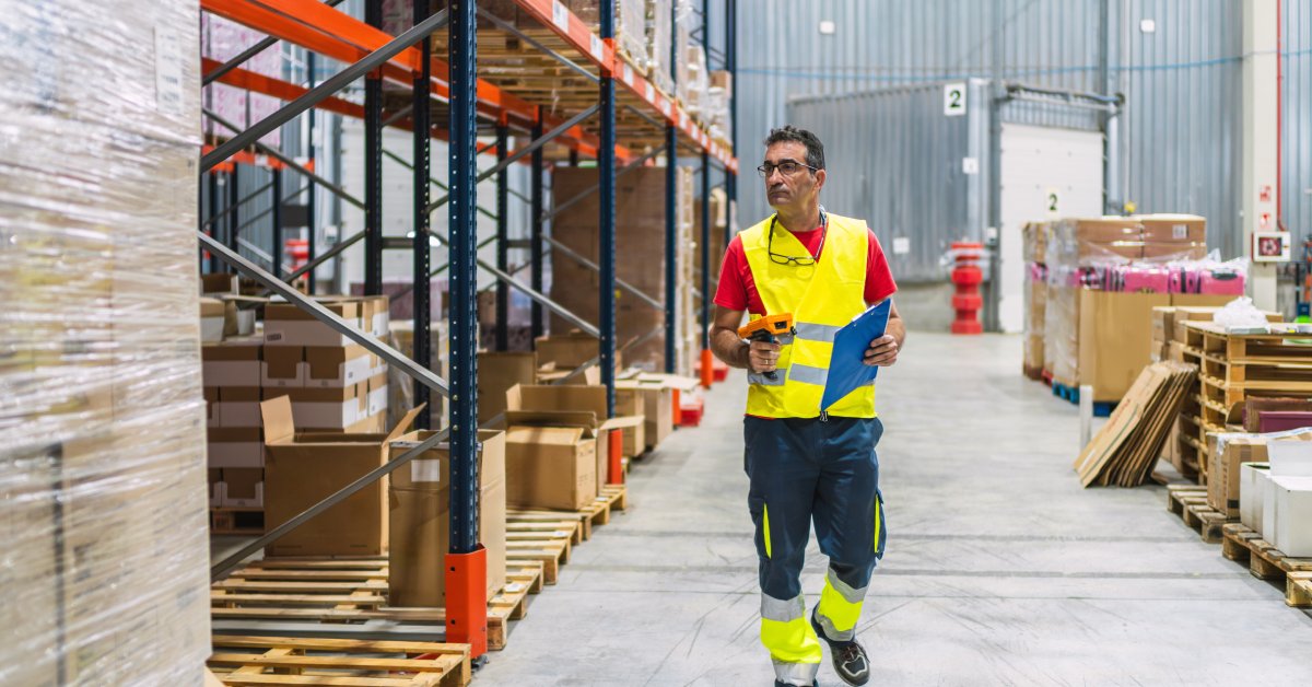 A warehouse worker wearing reflective gear, holding a scanning tool and a clipboard. He walks down an aisle.