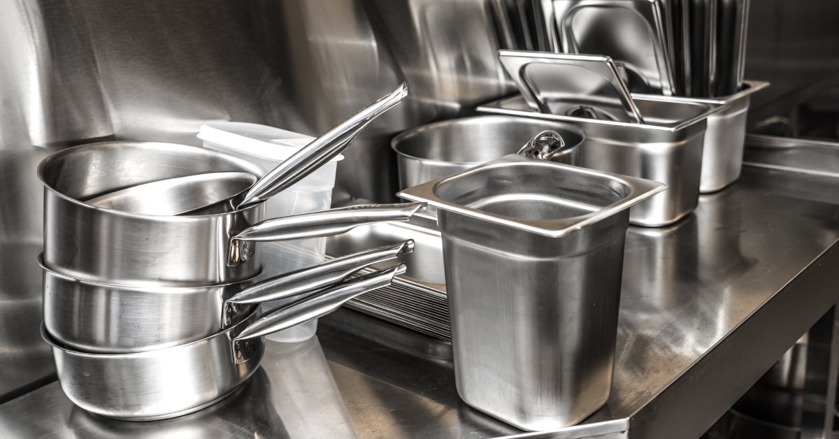 Aluminum kitchen tools sitting on a reflective counter. There are pots, trays, and containers stacked on the counter.