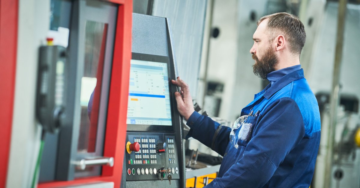 An industrial worker operating a manufacturing machine. He is looking at a large monitor with multiple buttons underneath.